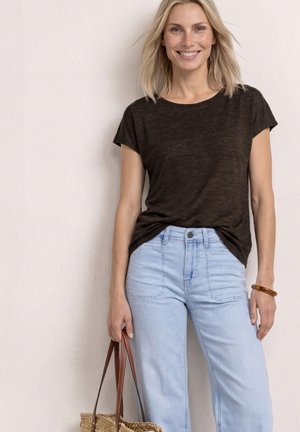 Smilende kvinde med blondt hår har en mørkebrun kortærmet top på, lyseblå jeans og holder en flettet taske med brune læderstropper.