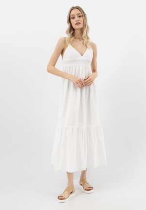 Donna bionda in piedi che indossa un abito maxi bianco senza maniche con vita arricciata e sandali beige con zeppa su sfondo bianco.