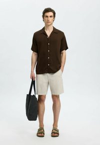 Kortärmad brun skjorta med knappar, randiga ljusa shorts, svart toteväska och bruna sandaler, stående mot en neutral bakgrund.