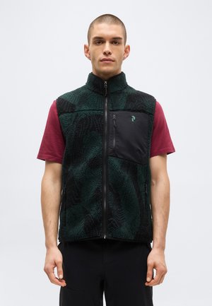 Férfi sötétzöld és fekete polár mellényben, cipzáras mellzsebbel, rövid ujjú bordó ing fölött, egy egyszerű háttér előtt áll.