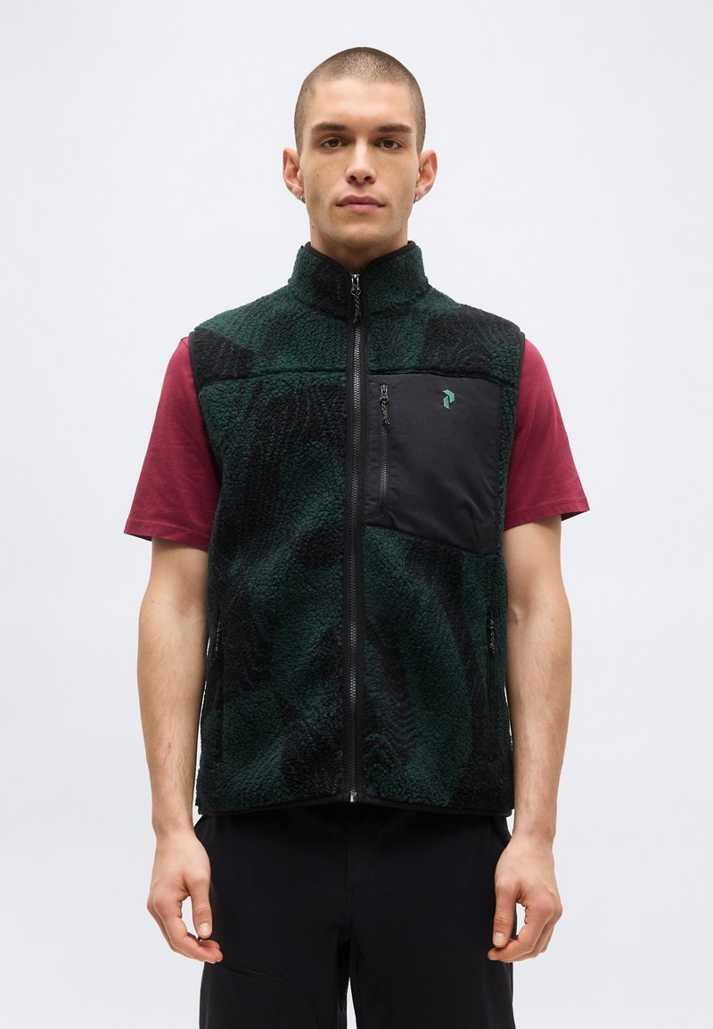 Άνδρας που φορά σκούφο πράσινο και μαύρο φλις γιλέκο με τσέπη στο στήθος με φερμουάρ πάνω από ένα κοντομάνικο μπορντό πουκάμισο, στέκεται σε απλό φόντο.