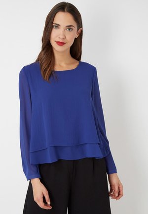 Blusa de manga larga azul real con un diseño en capas, una textura suave y un escote redondo. Combinada con pantalones negros.