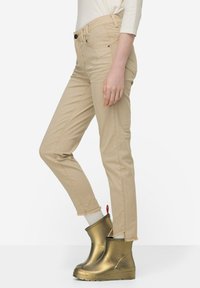 Béžové upnuté kalhoty s vysokým pasem, s bočními švy a třepeným lemem. Spárované s lesklými zlatými kotníkovými botami a bílými ponožkami.