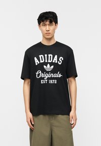 Νέος άνδρας με μαύρα κυματιστά μαλλιά που φοράει μαύρο T-shirt Adidas Originals και χακί παντελόνι, στέκεται μπροστά σε απλό λευκό φόντο.