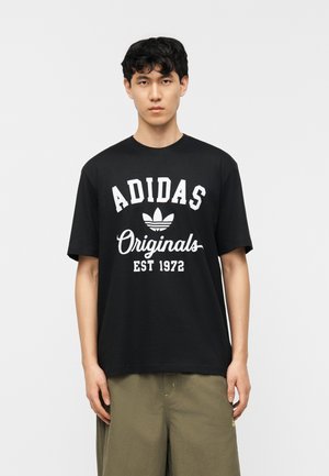 Νέος άνδρας με μαύρα κυματιστά μαλλιά που φοράει μαύρο T-shirt Adidas Originals και χακί παντελόνι, στέκεται μπροστά σε απλό λευκό φόντο.