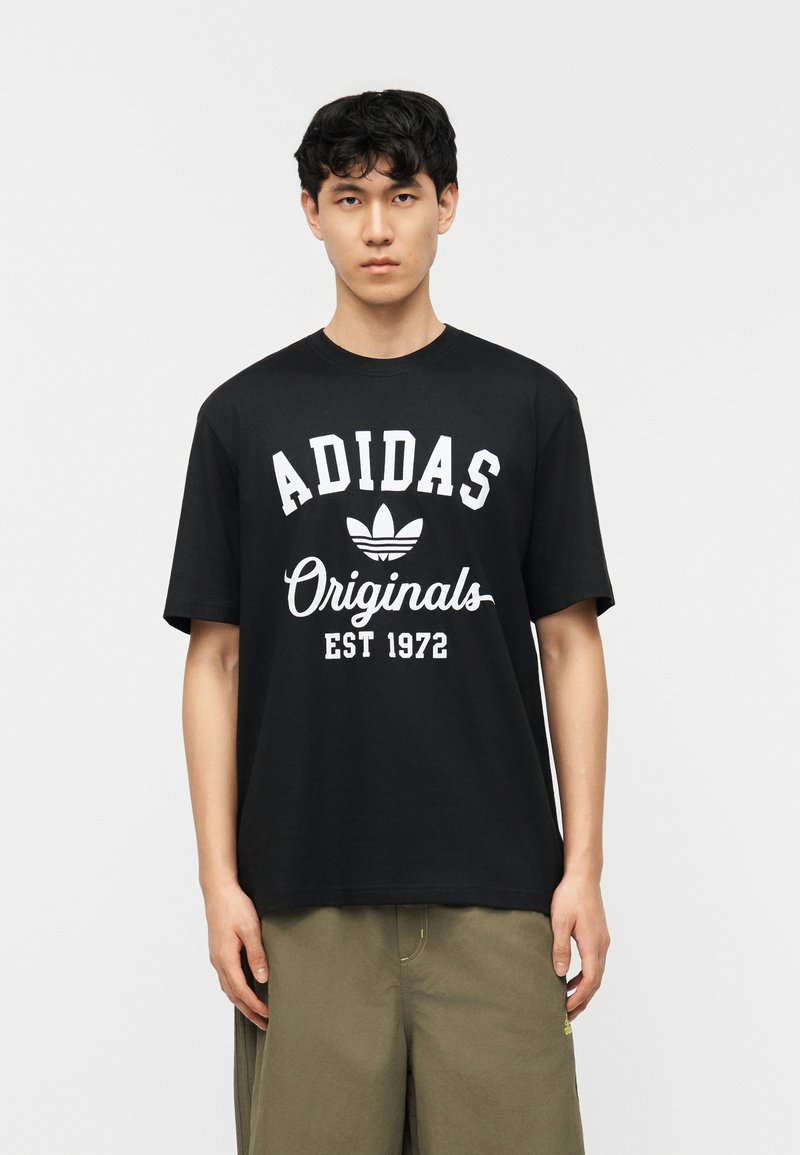 Νέος άνδρας με μαύρα κυματιστά μαλλιά που φοράει μαύρο T-shirt Adidas Originals και χακί παντελόνι, στέκεται μπροστά σε απλό λευκό φόντο.