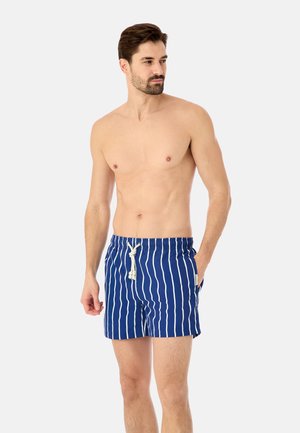 Oberkörperfreier Mann trägt blaue Badeshorts mit vertikalen weißen Streifen und gebundenem weißen Kordelzug, steht mit einer Hand in der Tasche.