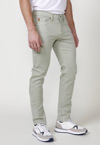 Jeans ajustados de color verde claro, fabricados en suave denim, con cinco bolsillos y detalles en hardware marrón. Se llevan con zapatillas deportivas blancas y grises.