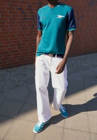 Polo à manches courtes bleu sarcelle et bleu marine avec logo brodé, associé à un pantalon blanc à jambes larges et des baskets bleu sarcelle avec des accents blancs.