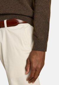Close-up of person wearing cream trousers, brown leather belt, and brown sweater with visible hand resting near the pocket.