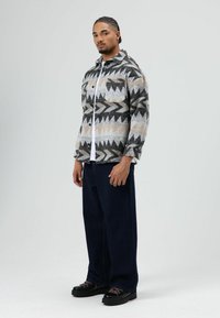 Wollen overhemd met patroon en geometrische ontwerpen in zwart, grijs en beige, met een knoopsluiting aan de voorkant, een borstzak en een losse pasvorm. Gecombineerd met donkere, wijd uitlopende broeken en stevige zwarte schoenen.