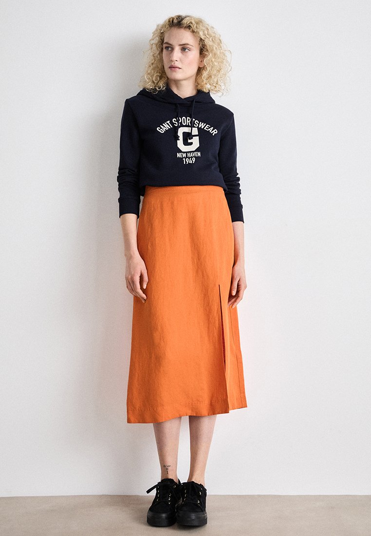 Marinblaues cropped Hoodie mit weißem Aufdruck, kombiniert mit einem orangefarbenen Leinen-Midirock mit einem seitlichen Schlitz. Schwarze Sneakers runden das Outfit ab.