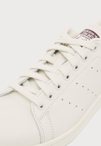 Бяла маратонка Adidas Stan Smith с бели връзки и перфорирани странични детайли на светъл фон.