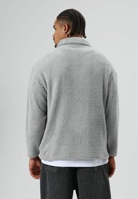Grijze fleece pullover met een zachte, gestructureerde oppervlakte. Voorzien van een hoge kraag en een losse pasvorm, gecombineerd met donkergrijze jeans.