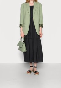 Blazer vert clair avec des poignets en satin roulés, robe noire à étages, sac à main vert et sandales noires à lanières avec une semelle naturelle.