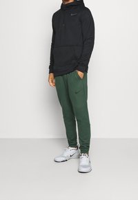 Homme portant un hoodie noir Nike, un jogging vert foncé Nike et des sneakers blanches Nike se tenant sur un fond léger uni.