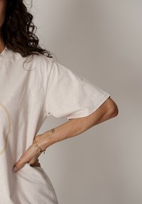 Jasnobeżowa oversize koszulka z okrągłym dekoltem i krótkimi rękawami. Widoczna miękka faktura. Ręka na talii, ozdobiona złotą bransoletką.