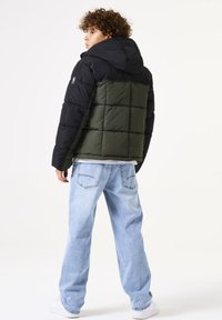 Veste matelassée avec capuche et manches noires, un corps vert olive et un design matelassé. Associée à un jean bleu clair et des baskets blanches.