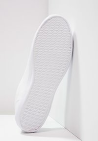 Semelle de basket blanche avec un motif de bande de roulement ondulé, positionnée verticalement contre un mur de coin blanc et un sol blanc.