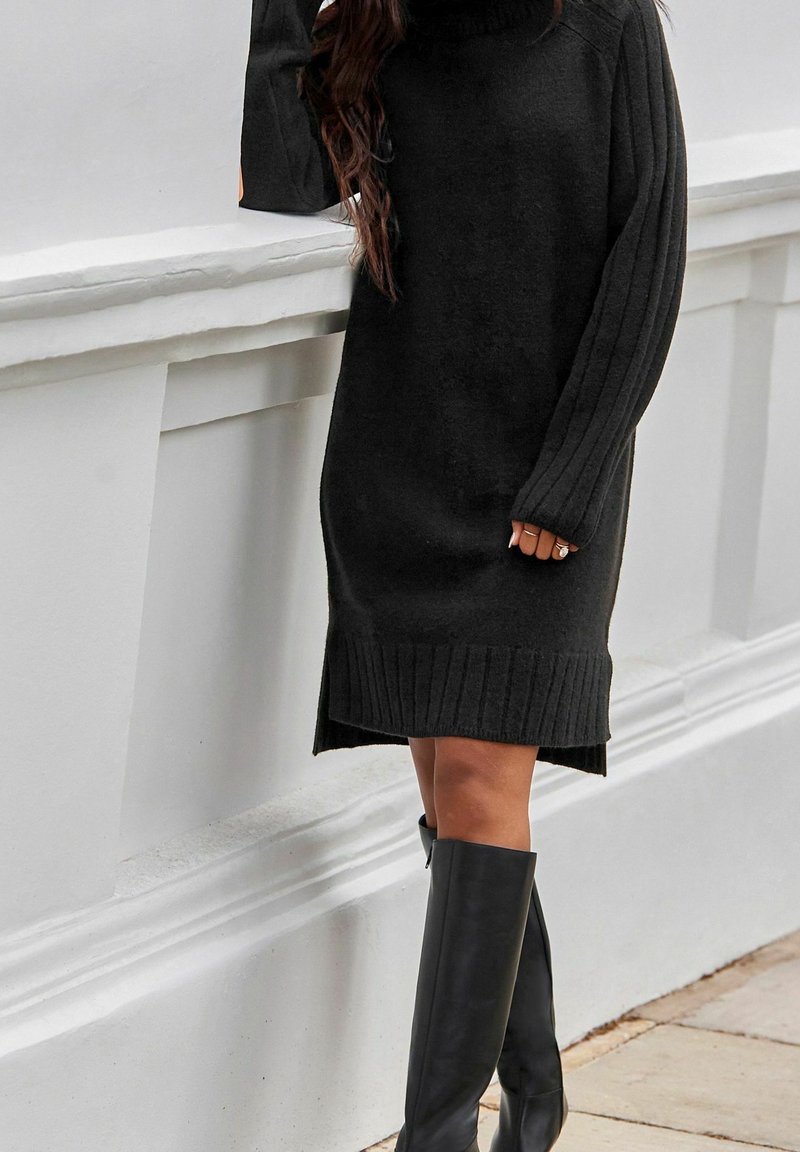 Femme portant une robe noire en tricot à manches longues et des bottes noires montantes, appuyée contre un mur blanc à l'extérieur.