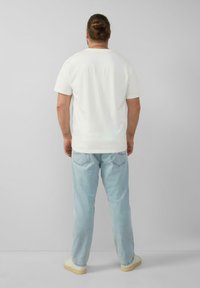 Man met een bun-kapsel, gekleed in een eenvoudig wit T-shirt, lichtblauwe jeans en witte sneakers, staand tegen een effen lichtgrijze achtergrond.