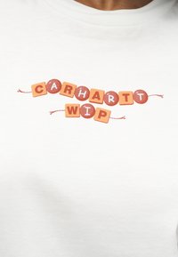 Λευκό πουκάμισο με πορτοκαλί και κόκκινα ραμμένα γράμματα που σχηματίζουν τη λέξη «CARHARTT WIP», τοποθετημένα σαν χάντρες σε δύο κορδόνια κατά μήκος του στήθους.