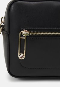 Borsa a tracolla nera in pelle con una cerniera dorata e un accento decorativo a forma di spilla da balia. Finitura testurizzata con una forma rettangolare elegante.