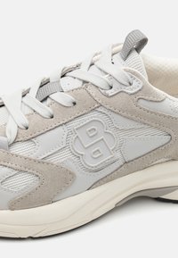 Detailní záběr na béžovou a šedou tenisku s texturovanými semišovými a síťovanými panely, bílými tkaničkami a vyvýšeným logem "BB" na boku.