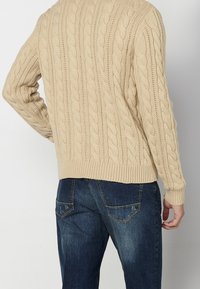 Pull beige en maille torsadée avec un motif texturé, ourlet et poignets côtelés, associé à un jean denim bleu foncé avec poches arrière.