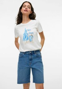 Camiseta blanca con gráfico azul de una sombrilla y una silla de playa, texto "Colores de Grecia". Combinada con pantalones cortos de mezclilla azul de longitud media.