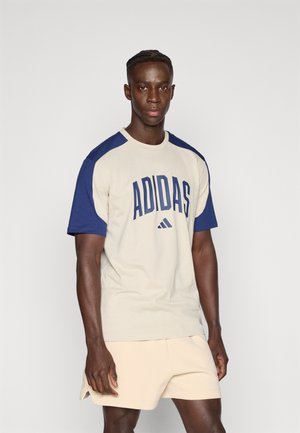 Camiseta beige con mangas raglán azul marino, con un gran texto azul "ADIDAS". Combinada con pantalones cortos beige claro. Textura suave.
