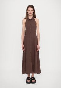 Brun lin halterkjole med knappefront, A-formet silhuett og kragedetalj. Modell iført kraftige svarte sandaler.