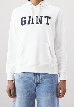 Personne portant un hoodie blanc avec le texte "GANT" et une poche avant, associé à un jean en denim bleu clair sur un fond uni.