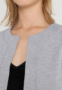 Femme portant un cardigan ouvert gris clair par-dessus un top noir à col en V, révélant le cou et la partie supérieure de la poitrine.