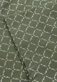 Zelená tkanina se opakujícím bílým geometrickým vzorem vzájemně propojených čtverců a kurzivním textem, s viditelnými detaily šití.