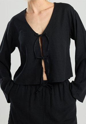 Top cropped negro de manga larga con tres lazos en la parte delantera, combinado con pantalones de cintura alta a juego, manos en los bolsillos, mostrado de cuello a caderas.