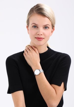 Silver mesh wristwatch with a white face and metallic hour markers, worn on a hand with fingers resting on the neck. Black short-sleeved top.