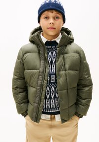 Doudoune verte avec capuche, boutons-pression et design matelassé. Portée par-dessus un sweat à motifs bleu marine et un pantalon beige.