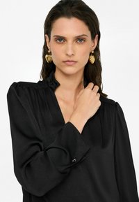 Blusa nera con finitura testurizzata, scollatura a V, maniche lunghe e dettagli arricciati sulle spalle. Gli orecchini sono dorati, a forma di cuore e metallici.