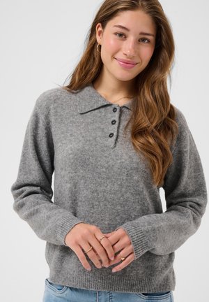 Jeune femme aux longs cheveux bruns porte un pull gris boutonné et des bagues en or, souriant doucement devant un fond clair uni.