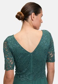 Robe teal avec un corsage en dentelle aux motifs floraux, manches courtes et un dos en V. Comprend une fermeture éclair dissimulée au dos.