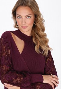 Burgundin värinen neulepusero, jossa on korkea kaulus. Puserossa on läpikuultavat pitsiset pitkät hihat, joissa on kukkakuviot, ja toisella puolella on aukko.