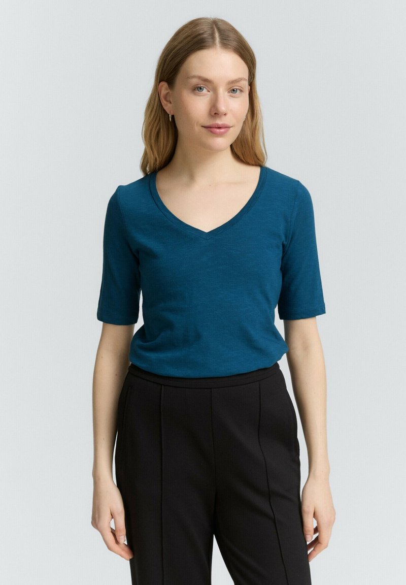 T-shirt verde acqua con scollo a V e maniche corte, realizzata in tessuto morbido, abbinata a pantaloni neri su misura, esposta su uno sfondo neutro.