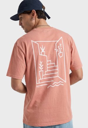 Hombre que lleva una camiseta color coral rosa con dibujo lineal blanco de escaleras, plantas y una puerta abierta en la parte trasera, combinado con una gorra azul marino.