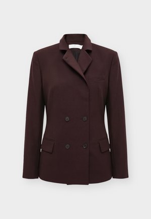 Mujer con cabello rubio corto viste un blazer marrón oscuro cruzado y pantalones rectos a juego con zapatos negros puntiagudos.