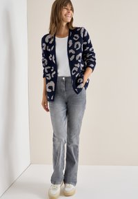 Námořnický svetr s jasně šedým leopardím potiskem, smíšené vlny, otevřený vpředu, kombinovaný se šedými džíny a bílými teniskami, s kapsami.