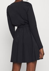 Robe noire à manches longues, corsage ajusté et jupe évasée. Tissu lisse avec une taille ceinturée et des boutons décoratifs sur les épaules.