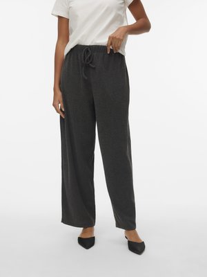 Άτομο που φοράει σκούρο γκρί φαρδιά παντελόνια με κορδόνι, λευκό κοντομάνικο πουκάμισο και μαύρα παπούτσια χωρίς κορδόνια με μυτερή μύτη, στέκεται σε απλό φόντο.