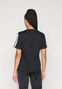 Haját copfba font nő áll háttal, fekete pólót és azonos színű nadrágot visel, fehér csíkokkal az ujjakon és az oldalakon.