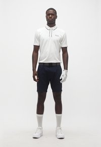 Άνδρας που φορά λευκό γάντι γκολφ, λευκό πόλο πουκάμισο, σκούρο μπλε σορτς, λευκές κάλτσες και λευκά αθλητικά παπούτσια, στέκεται μπροστά σε απλό λευκό φόντο.